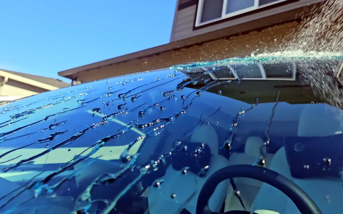 Close up of a garden hose spraying a windshield, with water beads and streaks running off the glass