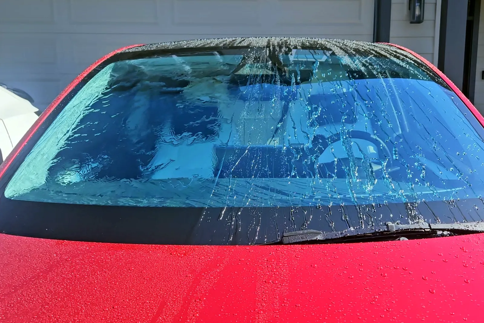 Red car windshield sprayed with a garden hose. The left half, untreated, is covered in a sheet of water. The right half, treated, has water running off in streaks and beads.